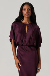 A woman is wearing a deep burgundy satin dress with a loose, draped bodice and short, wide sleeves. The dress features a small keyhole opening at the neckline. The fabric has a shiny, smooth texture.